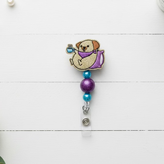 A novelty retractable badge reel featuring a glittery cartoon dog wearing a purple shirt and holding a blue cup, attached to blue and purple beads and a silver clip on a white background.