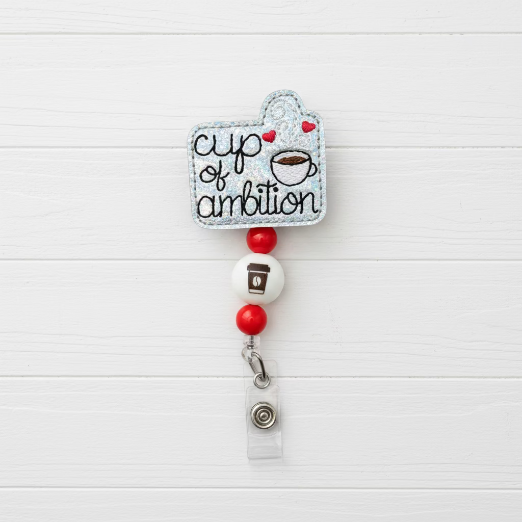 A novelty retractable badge reel featuring a glittery silver cup-shaped charm with the words 'cup of ambition' and a coffee cup graphic, accented with red and white beads and a clear plastic ID holder at the bottom.