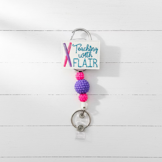 A custom retractable badge reel keychain featuring a white leather patch with the text 'Teaching with FLAIR' and a pink and purple 'X' design. The patch is attached to a purple and pink beaded cord, a silver metal carabiner, and a clear plastic badge reel on a light pink background.
