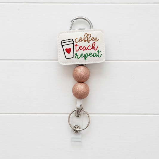 A coffee-themed teacher badge reel keychain with a square cream fabric tag reading 'coffee teach repeat' in brown, red, and green lettering, alongside a coffee cup graphic. The tag is attached to a silver carabiner, two glittery rose-gold beads, and a silver key ring with a clear plastic clip, shown on a light lavender background.