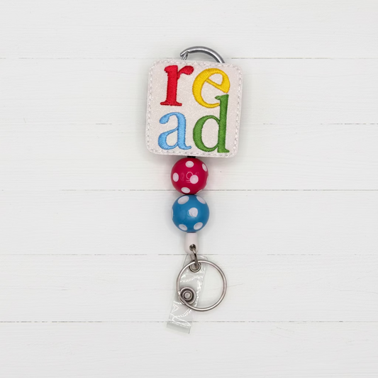 A retractable badge reel keychain featuring the word 'read' in colorful letters on a white faux leather patch, with two beaded spheres and a silver metal split ring on a light lavender background.