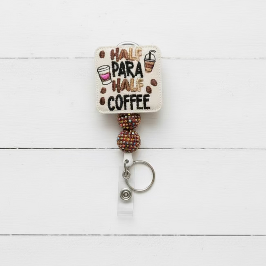 A retractable badge reel keychain with a square cream-colored embroidered patch that reads 'HALF PARA HALF COFFEE' in black and brown lettering, decorated with coffee cup and bean illustrations. The patch is attached to two dark purple beaded balls and a silver metal key ring on a light pink background.