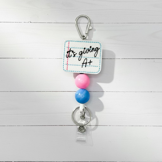 A novelty retractable badge reel keychain featuring a square white tag with a lined notebook paper design and the handwritten text 'it's giving A+'. The tag is attached to a silver metal carabiner, a pink and blue bead, and a white cord with a clear plastic badge holder, all set against a light lavender background.