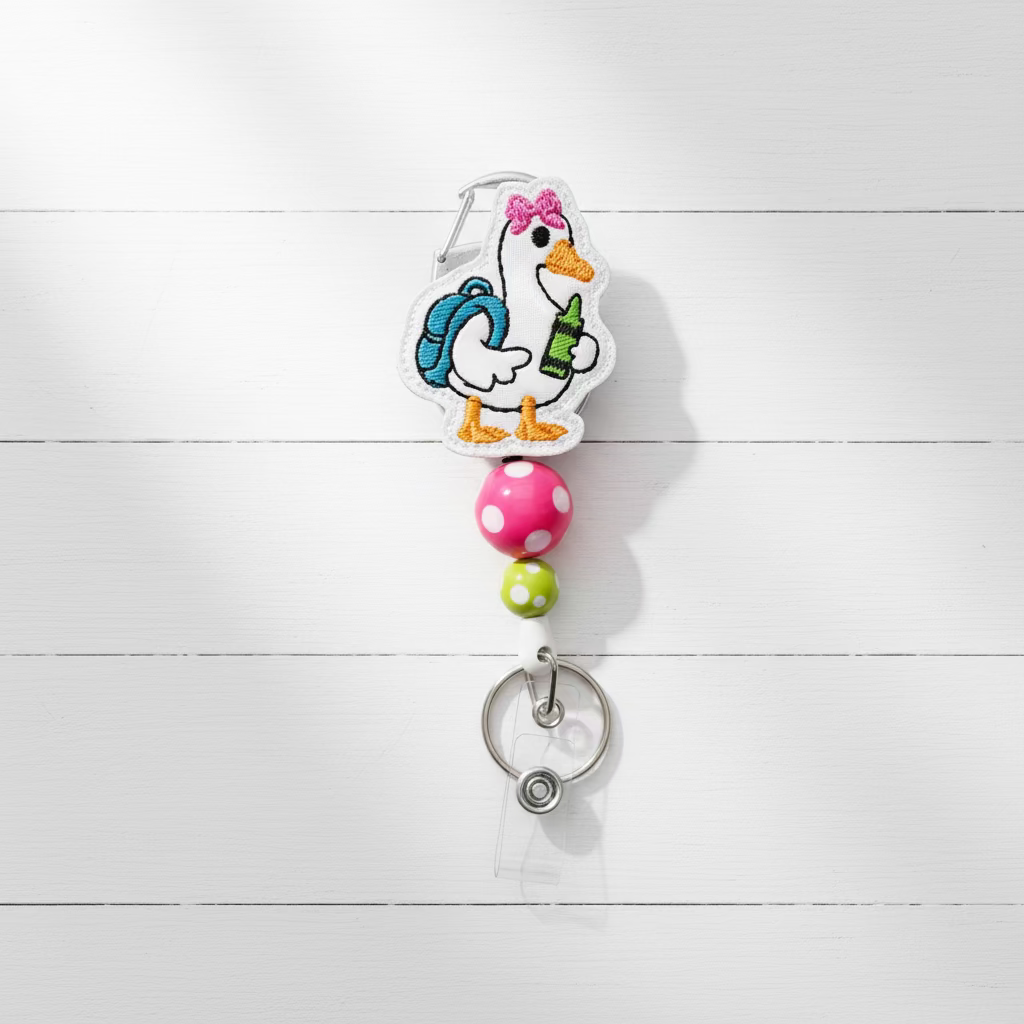 A cartoon duck badge reel keychain featuring a white duck with a blue backpack, pink bow, and green bottle, attached to a pink and green beaded strap with a silver metal clip on a light purple background.