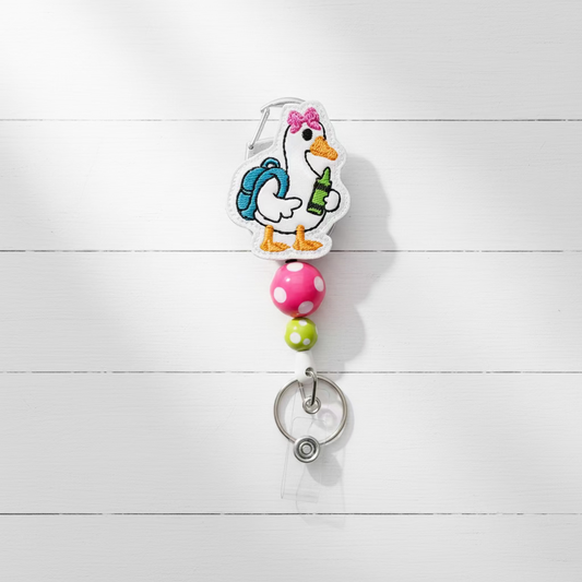 A cartoon duck badge reel keychain featuring a white duck with a blue backpack, pink bow, and green bottle, attached to a pink and green beaded strap with a silver metal clip on a light purple background.