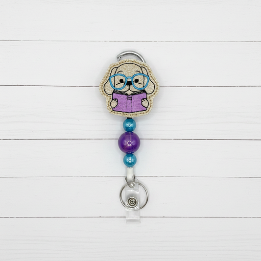 A novelty retractable badge reel featuring a cartoon elderly woman with glasses reading a purple book, attached to a silver metal clip and a clear plastic reel holder on a light lavender background.