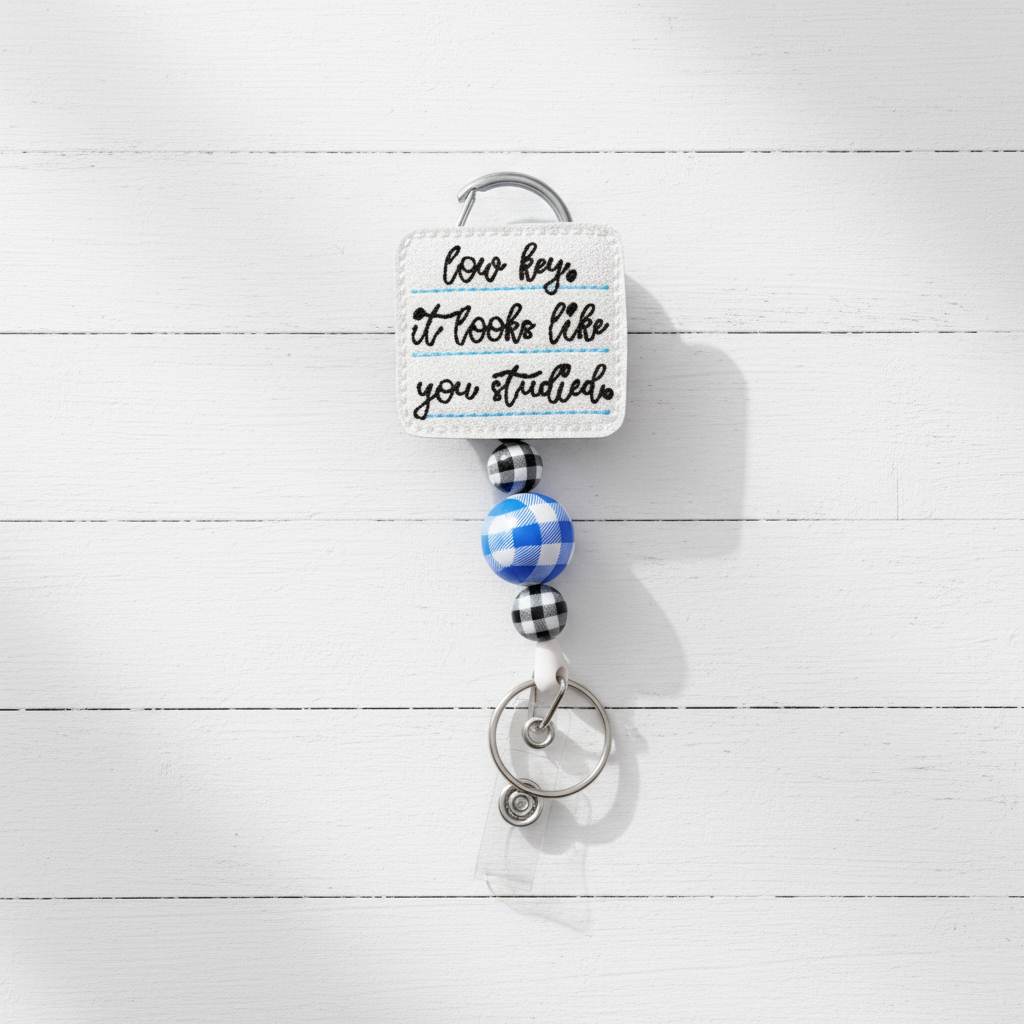 A novelty keychain with a square white embroidered patch reading 'how key it looks like you studied' in black script, attached to a silver carabiner and key ring with blue and black gingham beads on a light lavender background.