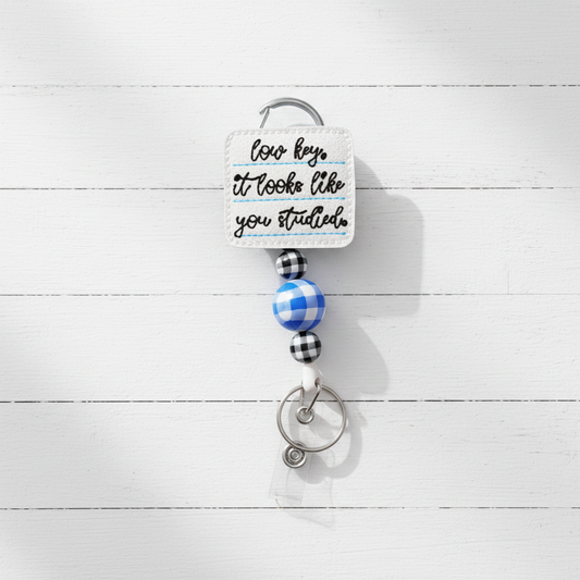 A novelty keychain with a square white embroidered patch reading 'how key it looks like you studied' in black script, attached to a silver carabiner and key ring with blue and black gingham beads on a light lavender background.