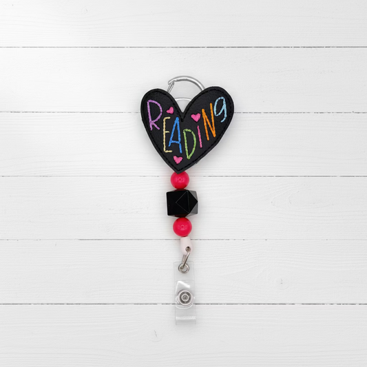 A heart-shaped black chalkboard-style badge reel with the word 'READING' in colorful letters, attached to red beads, a black faceted bead, and a clear plastic ID holder on a silver key ring, photographed on a light pink background.