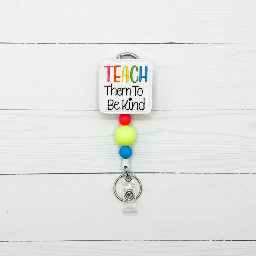 A self-adhesive square patch with the text 'TEACH Them To Be Kind' in colorful and black lettering, attached to a silver carabiner and a retractable badge reel with colorful beads.