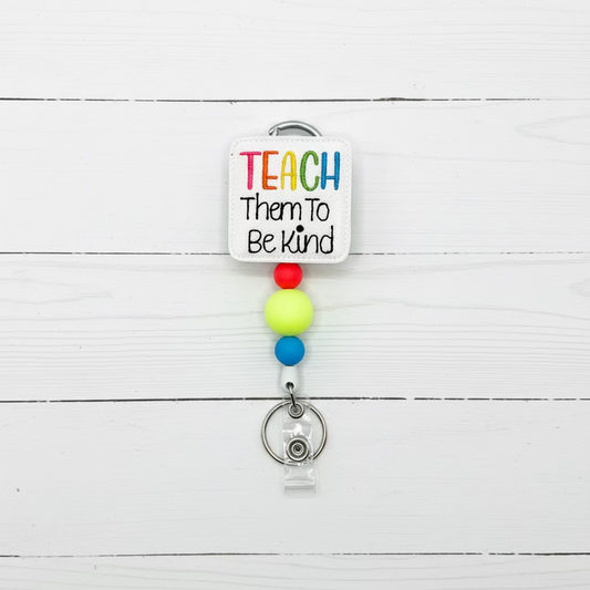 A self-adhesive square patch with the text 'TEACH Them To Be Kind' in colorful and black lettering, attached to a silver carabiner and a retractable badge reel with colorful beads.
