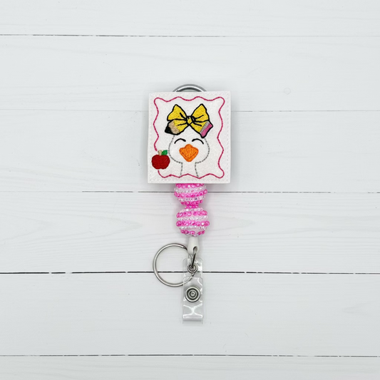A pink and white retractable badge reel keychain featuring a cartoon penguin wearing a yellow bow and holding a red apple, with a silver key ring and clear plastic ID holder.