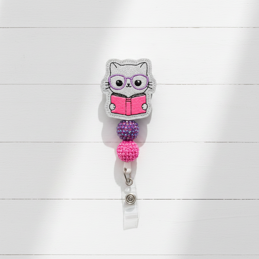 A rhinestone-covered retractable badge reel keychain featuring a cartoon owl wearing purple glasses and reading a pink book, with purple and pink beaded sections and a silver key ring on a light lavender background.