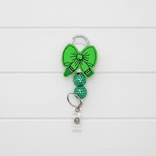 A bright green felt bow keychain accessory with a silver metal carabiner clip, a beaded green chain section, and a clear plastic badge holder at the bottom, shown on a light pink background.