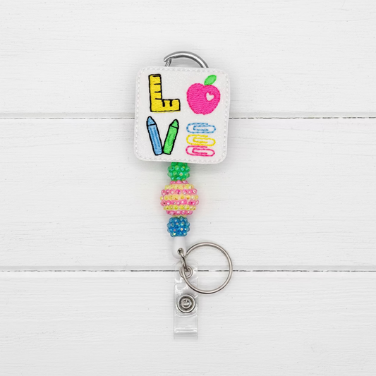 A square white leatherette badge reel keychain featuring colorful embroidered school-themed letters and icons, including a pink apple, yellow ruler, blue and green pencils, and paperclips. The reel is attached to a silver metal key ring and a clear plastic ID tag, set against a solid pale lavender background.