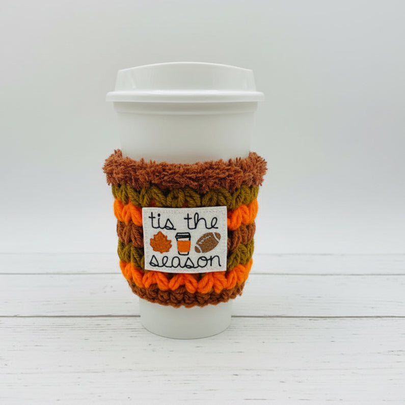 A white paper coffee cup with a lid, wrapped in a crocheted autumn-themed sleeve. The sleeve features stripes of brown, orange, and olive green, with a white label that reads 'tis the season' decorated with small fall icons.