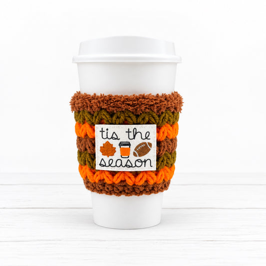 A white paper coffee cup with a lid, wrapped in a crocheted autumn-themed sleeve. The sleeve features stripes of brown, orange, and olive green, with a white label that reads 'tis the season' decorated with small fall icons.