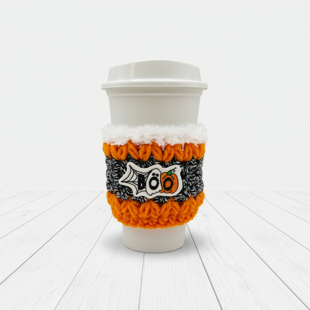 A white paper coffee cup with a white lid, wrapped in a knitted Halloween-themed sleeve featuring orange, black, and white colors with a '100' pumpkin design, on a light gray background.