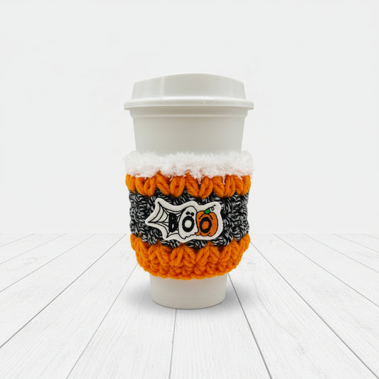 A white paper coffee cup with a white lid, wrapped in a knitted Halloween-themed sleeve featuring orange, black, and white colors with a '100' pumpkin design, on a light gray background.