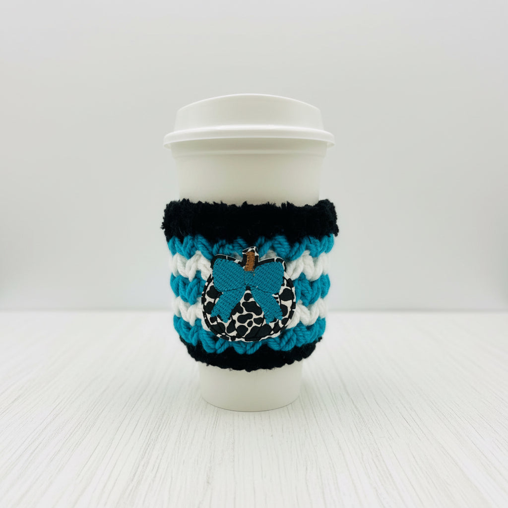 A white paper coffee cup with a white lid, wrapped in a black and white striped knit sleeve featuring a blue bow applique with a leopard print center and a small gold charm.