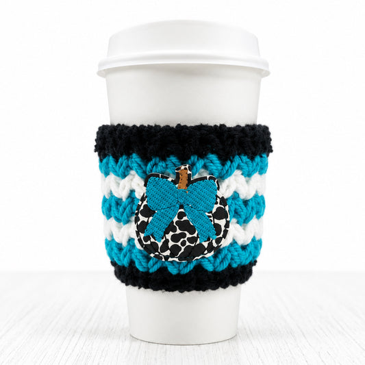 A white paper coffee cup with a white lid, wrapped in a black and white striped knit sleeve featuring a blue bow applique with a leopard print center and a small gold charm.