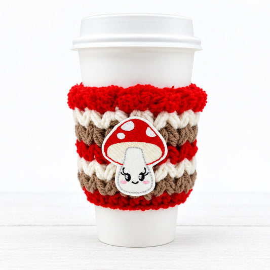 A white reusable coffee cup with a red, cream, and brown knitted sleeve featuring a cute embroidered mushroom patch.