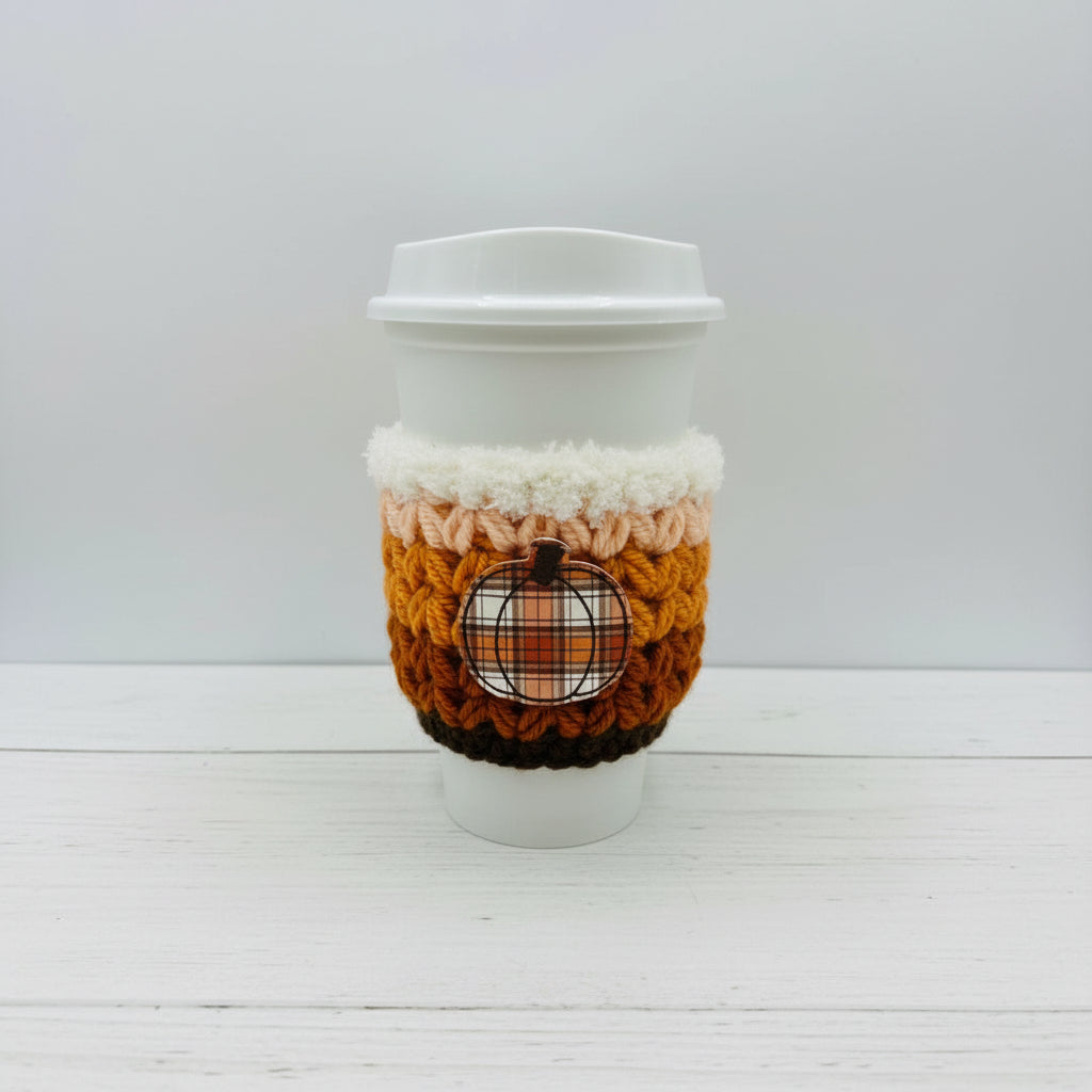 A white paper coffee cup with a white lid, wrapped in a crocheted autumn-themed sleeve featuring stripes of cream, peach, orange, and brown, with a small plaid pumpkin patch detail on the front.