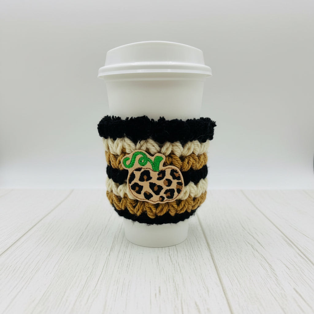 A white paper coffee cup with a white lid, wrapped in a black and tan crochet sleeve featuring a leopard-print pumpkin patch and a green embroidered 'SO' monogram, on a light gray background.
