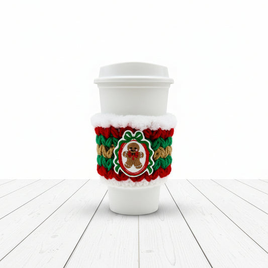 A white disposable coffee cup with a white lid, wrapped in a red, green, and white crocheted holiday sleeve featuring a gingerbread man patch in the center, on a light gray background.