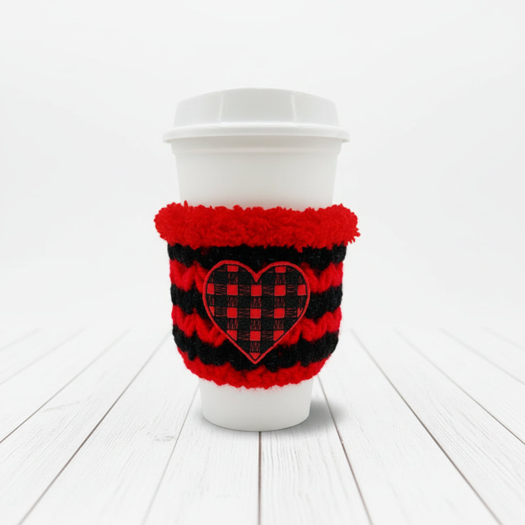 A white paper coffee cup with a white lid, wrapped in a red and black knit sleeve featuring a red and black checkered heart design, set against a plain light gray background.