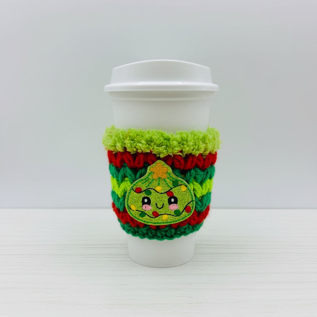 A white paper coffee cup with a white lid, wrapped in a bright green, red, and green crocheted holiday cozie featuring a cute embroidered Christmas tree face.