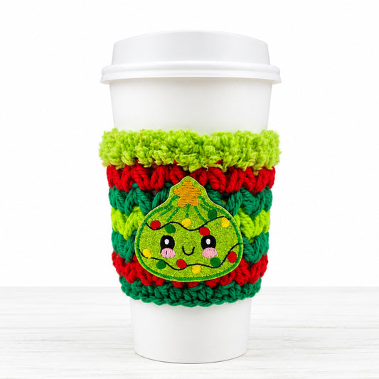 A white paper coffee cup with a white lid, wrapped in a bright green, red, and green crocheted holiday cozie featuring a cute embroidered Christmas tree face.