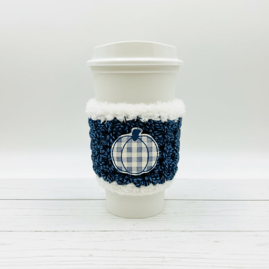 A white disposable coffee cup with a navy blue and white gingham knit sleeve featuring a faux-fur trim and a gingham pumpkin applique, set against a plain light background.