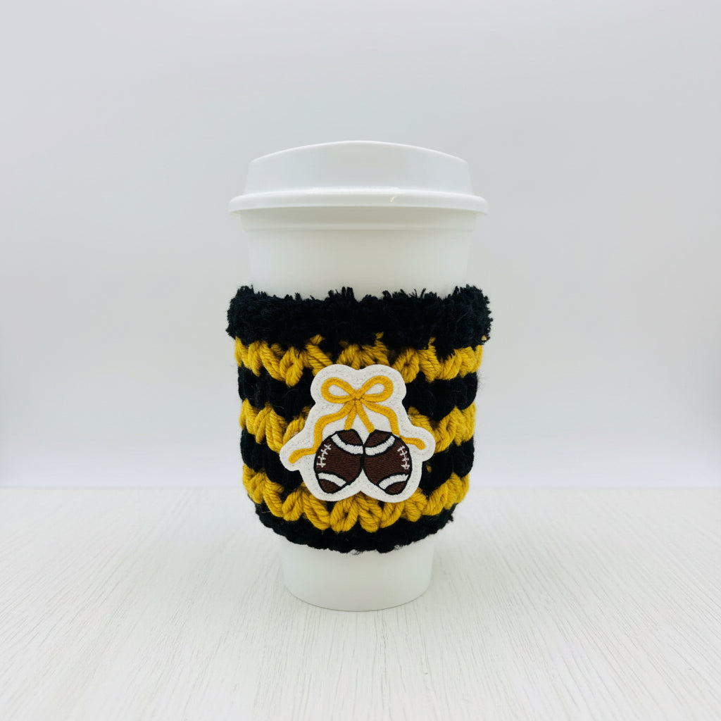 A white paper coffee cup with a black and yellow striped knit sleeve featuring a white bow and brown football patch, shown on a light gray background.