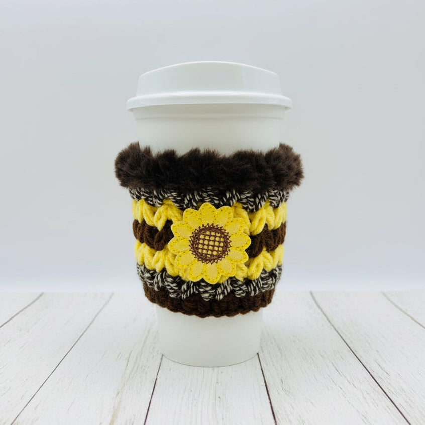 A white paper coffee cup with a white lid, wrapped in a handmade knit sunflower-themed sleeve featuring yellow and brown stripes, a central sunflower motif, and dark brown faux fur trim at the top, shown against a plain white background.