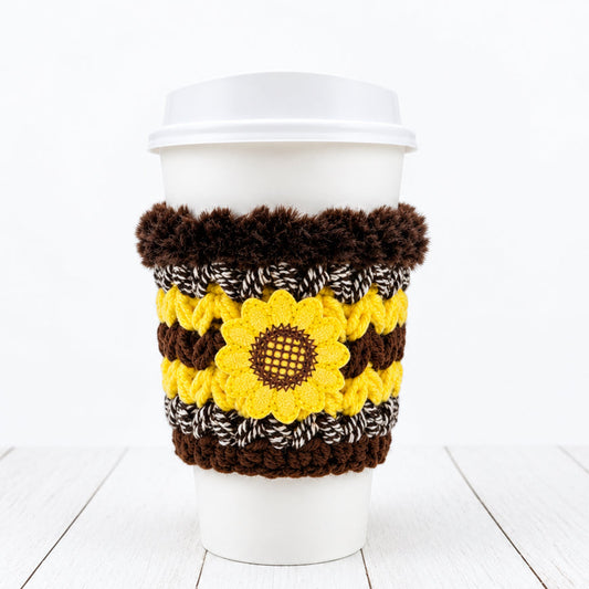 A white paper coffee cup with a white lid, wrapped in a handmade knit sunflower-themed sleeve featuring yellow and brown stripes, a central sunflower motif, and dark brown faux fur trim at the top, shown against a plain white background.