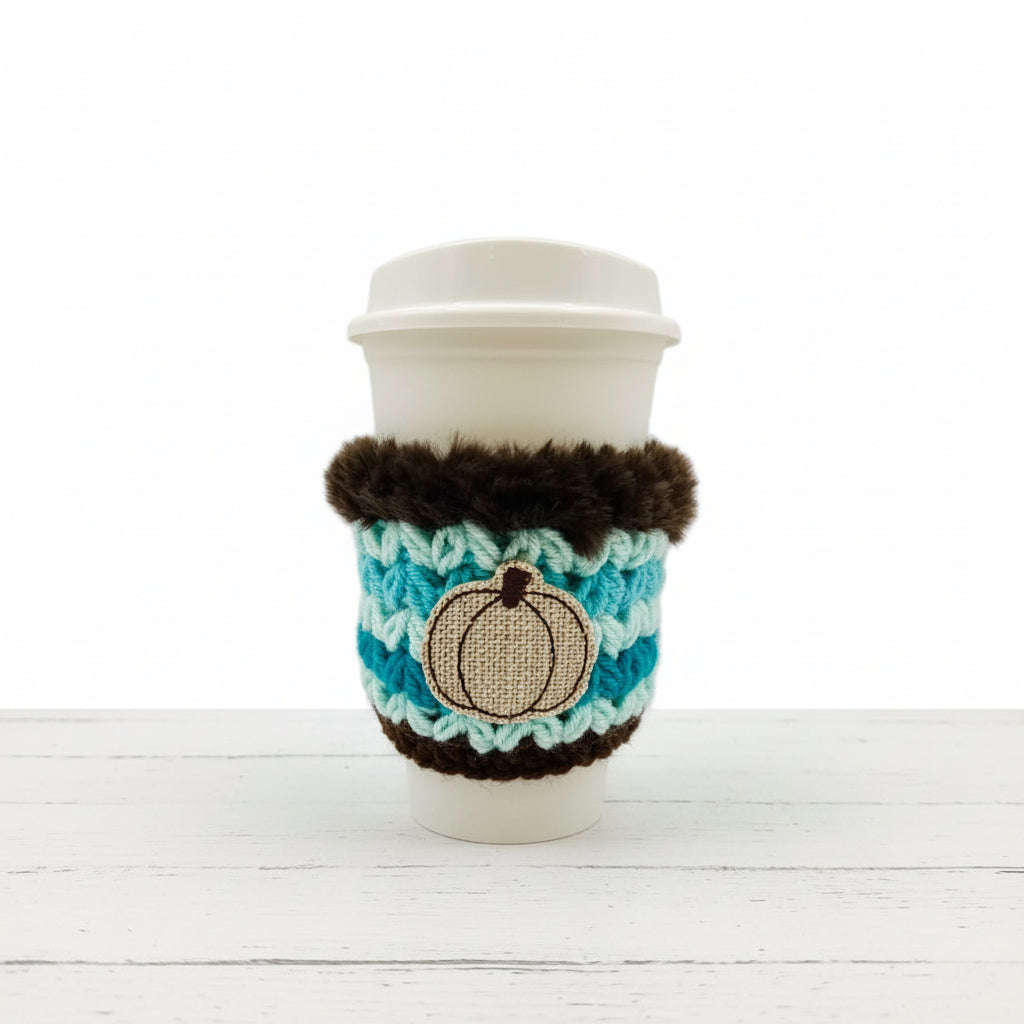 A white disposable coffee cup with a lid, wrapped in a turquoise and brown crocheted sleeve featuring a beige pumpkin applique, on a plain light background.