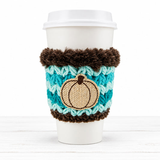 A white disposable coffee cup with a lid, wrapped in a turquoise and brown crocheted sleeve featuring a beige pumpkin applique, on a plain light background.