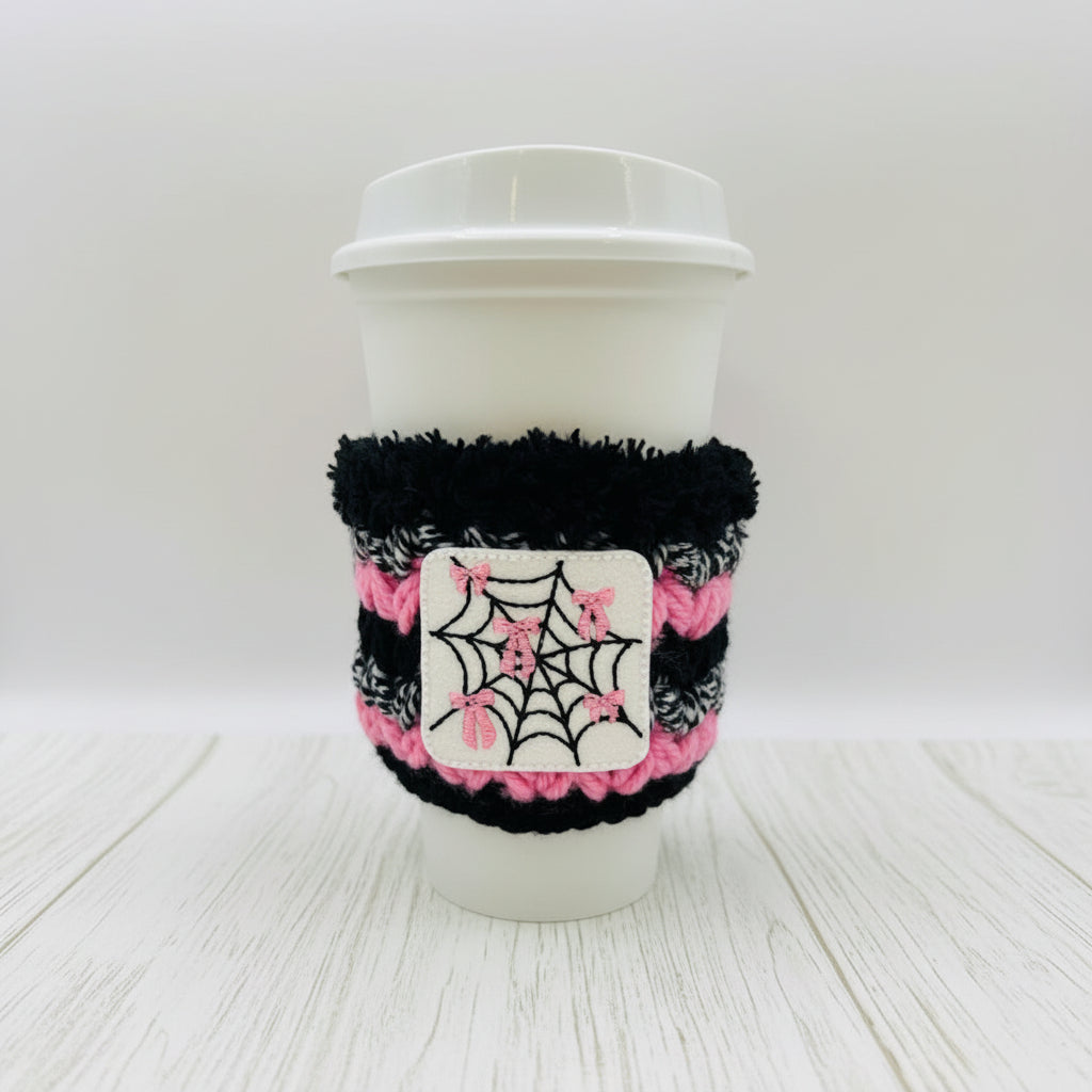 A white disposable coffee cup with a black and pink fuzzy sleeve featuring a white patch with a black spiderweb and pink bows, on a light gray background.