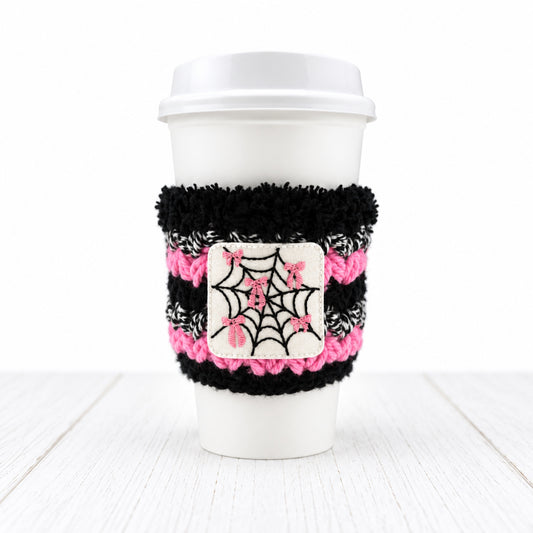 A white disposable coffee cup with a black and pink fuzzy sleeve featuring a white patch with a black spiderweb and pink bows, on a light gray background.