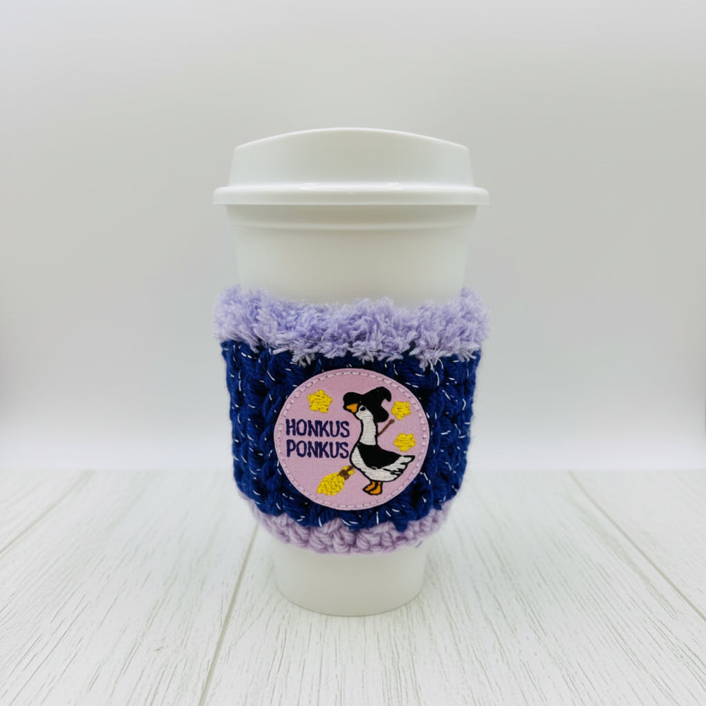 A white paper coffee cup with a white lid, wrapped in a blue and lavender knit sleeve featuring a circular pink patch with a penguin and the text 'HONKUS PONKUS'.