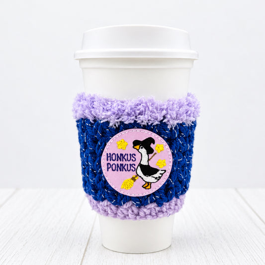 A white paper coffee cup with a white lid, wrapped in a blue and lavender knit sleeve featuring a circular pink patch with a penguin and the text 'HONKUS PONKUS'.