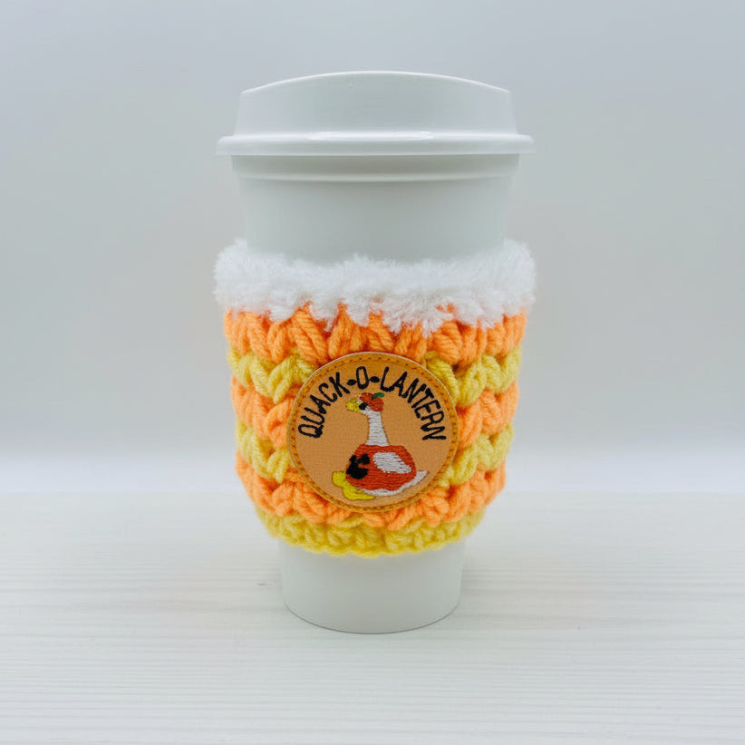 A white disposable coffee cup with a white lid, wrapped in a crocheted sleeve featuring orange, yellow, and white stripes, a faux-fur trim, and a circular 'Quack-O-Lantern' duck patch.