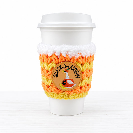 A white disposable coffee cup with a white lid, wrapped in a crocheted sleeve featuring orange, yellow, and white stripes, a faux-fur trim, and a circular 'Quack-O-Lantern' duck patch.