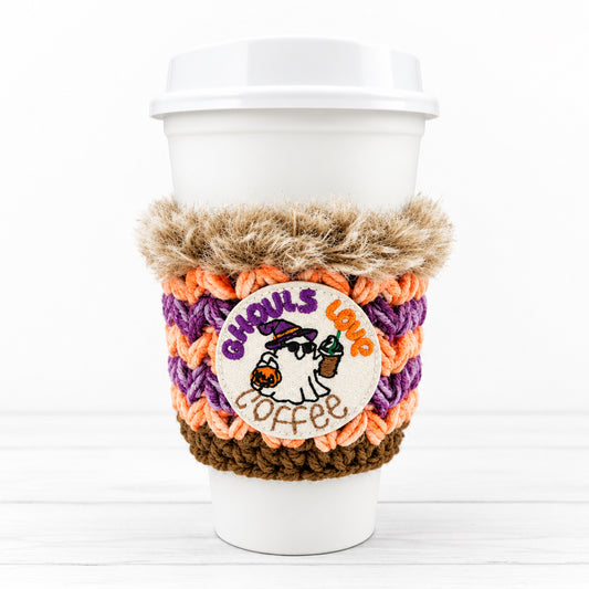 A white paper coffee cup with a lid, wrapped in a handmade crochet Halloween-themed coffee sleeve featuring orange, purple, brown, and beige colors, with a round embroidered 'Ghouls Lamp Coffee' patch.