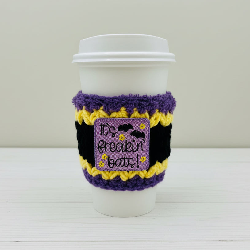 A white paper coffee cup sleeve with a purple fuzzy trim, black and yellow striped pattern, and a central purple patch that reads 'It's freakin' bats!' with bats and flowers.