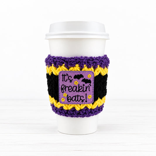 A white paper coffee cup sleeve with a purple fuzzy trim, black and yellow striped pattern, and a central purple patch that reads 'It's freakin' bats!' with bats and flowers.