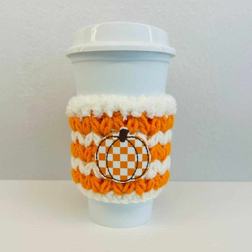 A white plastic takeaway coffee cup with a white lid, wrapped in an orange and white striped knit cozy featuring a central pumpkin design with a black and white checkered pattern.