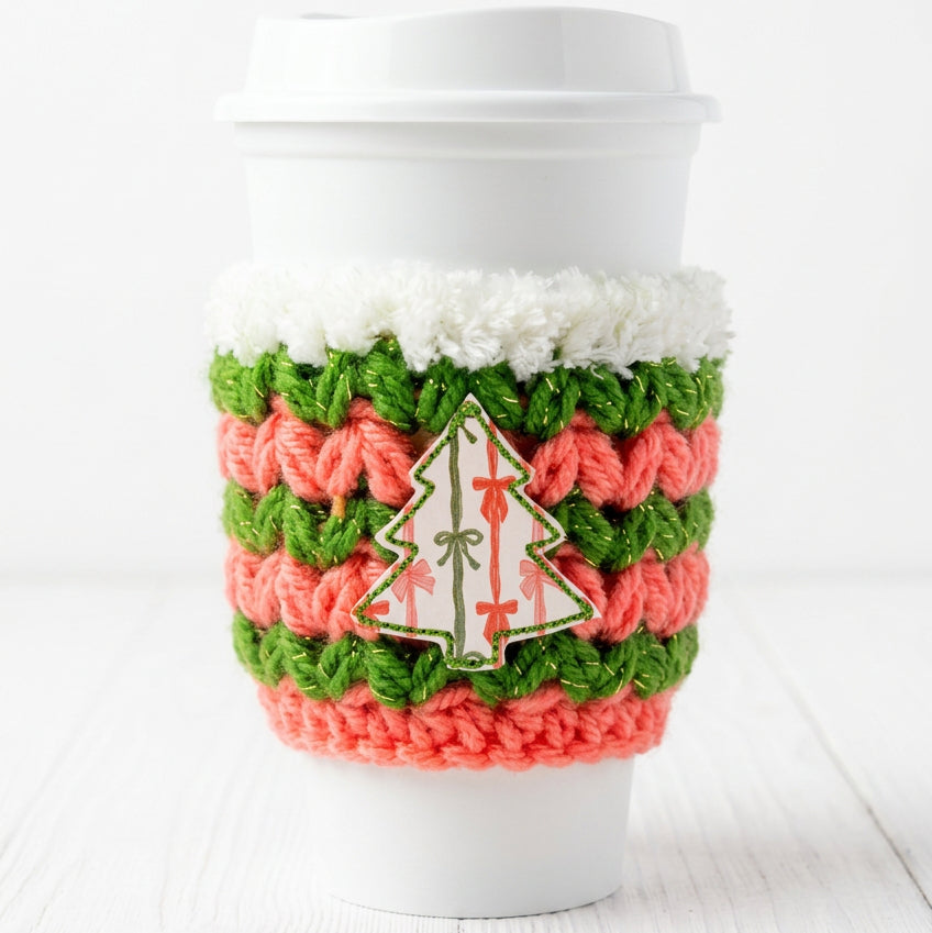 A white disposable coffee cup with a white lid, wrapped in a crocheted holiday kooshie featuring green and coral pink stripes, a white faux-fur trim, and a white Christmas tree patch with red ornaments.