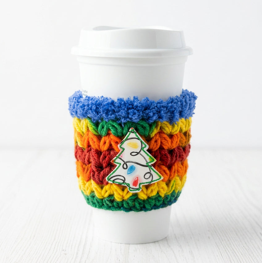 A white disposable coffee cup with a colorful crocheted sleeve featuring a Christmas tree patch, set against a plain light gray background.